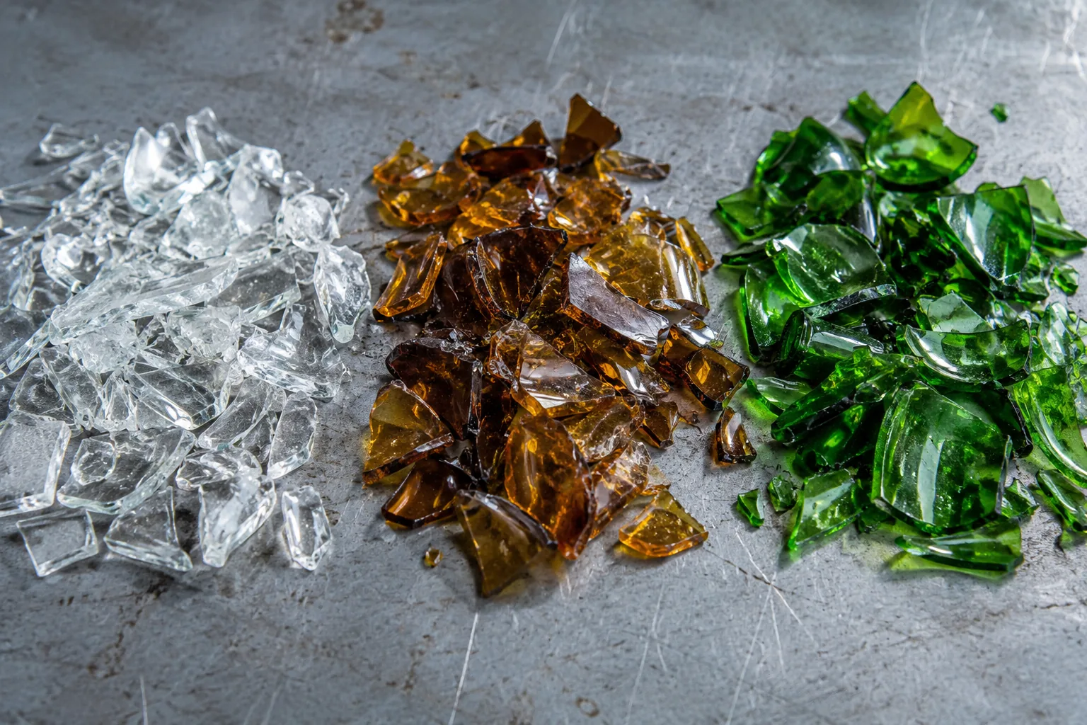Three separated piles of colorless, amber and green crushed bottle glass on a gray table